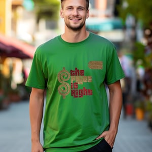 May include: A green short-sleeved t-shirt featuring a gold glitter dollar sign and the text "the price is right" in a dark red block font. The design includes a small gold glitter tag icon positioned above the text on the chest area.