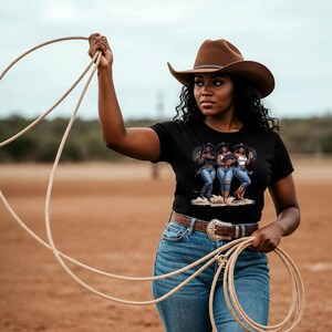 Op de afbeelding: Een vrouw draagt een bruine cowboyhoed en een zwart t-shirt met een afbeelding van drie vrouwen in cowboykleding. Ze houdt een touw vast en draagt een spijkerbroek en een bruine riem.