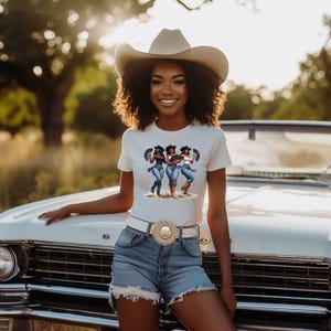 Op de afbeelding: Een vrouw draagt een wit t-shirt met een afbeelding van drie vrouwen in cowboyhoeden en jeans. Ze draagt ook een cowboyhoed, een spijkerbroek en een witte riem met een decoratieve gesp. De achtergrond omvat een witte auto en een natuurlijke omgeving.