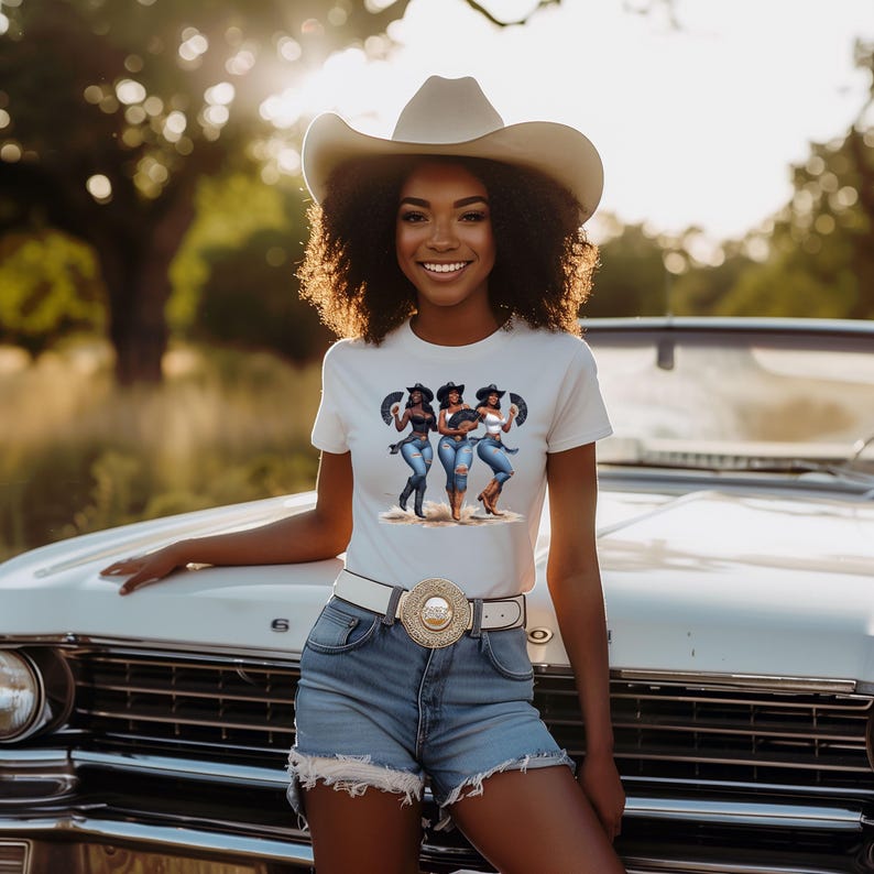 Op de afbeelding: Een vrouw draagt een wit t-shirt met een afbeelding van drie vrouwen in cowboyhoeden en jeans. Ze draagt ook een cowboyhoed, een spijkerbroek en een witte riem met een decoratieve gesp. De achtergrond omvat een witte auto en een natuurlijke omgeving.