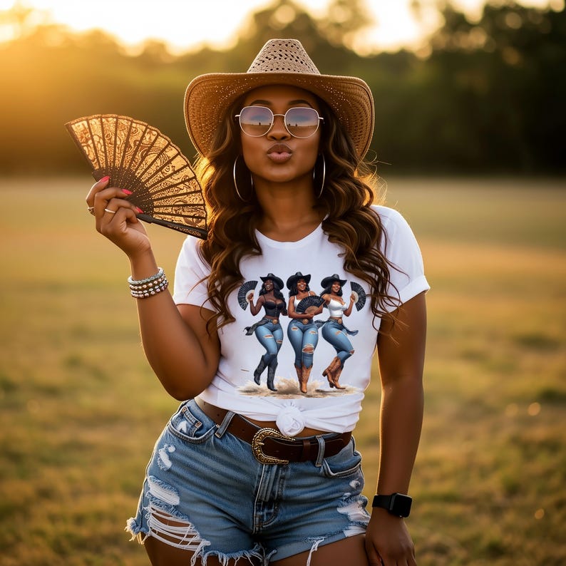 Op de afbeelding: Een vrouw draagt een strooien cowboyhoed, een zonnebril en een wit t-shirt met een afbeelding van drie vrouwen met cowboyhoeden. Ze houdt een decoratieve zwarte waaier vast en draagt een spijkerbroek met een bruine riem.