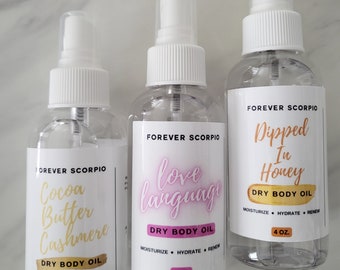May include: Three clear glass bottles with white spray tops and labels. The labels are white with black text and pink and gold accents. The bottles contain dry body oil with the scents Cocoa Butter Cashmere, Love Language, and Dipped in Honey. Each bottle is 4 oz.