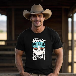 Puede incluir: Una camiseta negra con un estampado gr&aacute;fico de un rostro barbudo con el pelo trenzado que lleva gafas de sol que reflejan un paisaje des&eacute;rtico. Sobre el rostro aparece el texto Feelin' WILLIE y debajo la palabra Good en blanco.