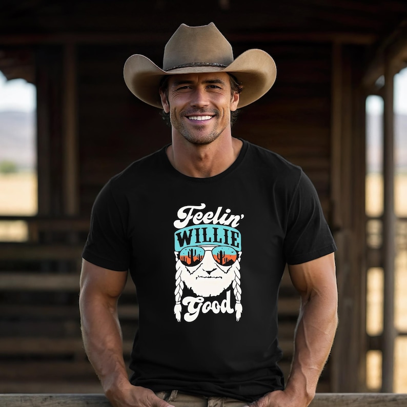 Puede incluir: Una camiseta negra con un estampado gr&aacute;fico de un rostro barbudo con el pelo trenzado que lleva gafas de sol que reflejan un paisaje des&eacute;rtico. Sobre el rostro aparece el texto Feelin' WILLIE y debajo la palabra Good en blanco.