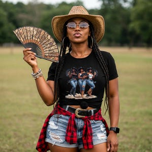Op de afbeelding: Een vrouw draagt een zwart t-shirt met een afbeelding van drie vrouwen in cowboyhoeden en jeans. Ze draagt ook een strohoed, zonnebril en houdt een decoratieve waaier vast. Ze draagt een rood-zwart geruit overhemd om haar middel geknoopt.