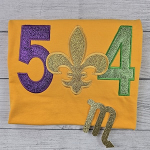 May include: Mustard yellow t-shirt with purple glitter number 5, a gold glitter fleur-de-lis, and green glitter number 4. A gold glitter letter "m" is below the numbers. The shirt is on a light wood grain surface.