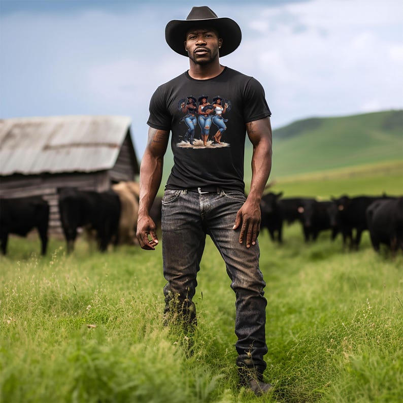 Op de afbeelding: Een zwart t-shirt met een afbeelding van drie vrouwen in spijkerbroeken en cowboyhoeden. De persoon draagt een zwarte cowboyhoed en een donkerblauwe spijkerbroek. De achtergrond omvat een schuur, koeien en een groen veld.