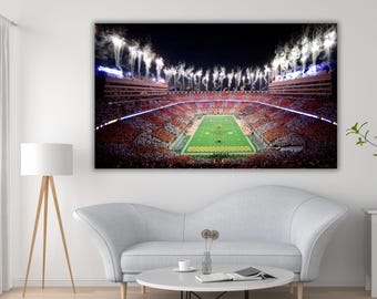 May include: A large rectangular wall art print depicting a night view of a football stadium filled with spectators. The field is illuminated in green, surrounded by orange seating sections, with white fireworks exploding above the stadium.