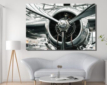 May include: A large wall art print featuring a close-up of a polished silver airplane propeller and engine cowling. The metallic surface reflects light, highlighting the circular engine housing and the long, tapered blades of the aircraft engine.