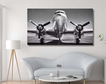 May include: A black and white wall art print featuring a front-facing view of a vintage twin-engine propeller airplane on a runway. The aircraft displays a metallic finish, two large propellers, and landing gear, captured in high contrast.
