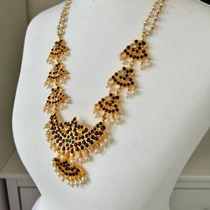 May include: A gold necklace featuring red and green gemstones and white pearls. The necklace has a large pendant in the centre and smaller pendants on either side.
