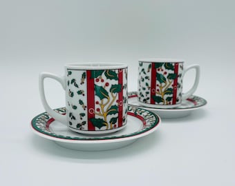May include: Two white ceramic coffee cups and saucers with a Christmas holly design. The cups and saucers have a red and green holly pattern with a white background. The cups have a handle.