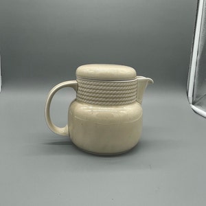 Peut inclure: Une crémier en céramique beige avec un motif en forme de corde autour du haut. La crémier a un couvercle et une poignée.