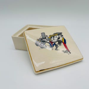 May include: Cream-colored square trinket box with a gold trim. The lid features a vintage car illustration with figures in colorful attire. The box is open, revealing its interior. A decorative piece for storing small items.