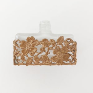 Puede incluir: Un frasco de perfume de vidrio esmerilado con un dise&ntilde;o floral de metal dorado alrededor de la parte superior. El dise&ntilde;o presenta flores y hojas delicadas.