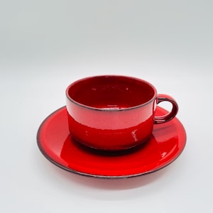 Puede incluir: Un vibrante juego de taza y platillo de cerámica roja. La taza tiene una forma redondeada con un asa pequeña, y el platillo es circular. Ambas piezas tienen un acabado brillante y un borde oscuro, creando un contraste llamativo.