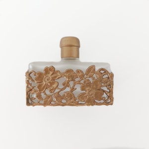 Puede incluir: Un frasco de perfume de vidrio esmerilado con un dise&ntilde;o floral dorado. El frasco tiene una tapa dorada y una forma rectangular.