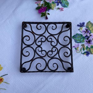 May include: A rustic, iron, square-shaped trivet with a decorative, swirling design. The trivet has four small, round feet.