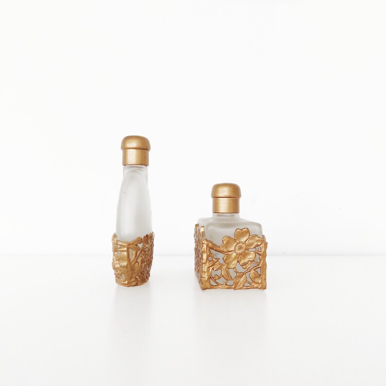Puede incluir: Dos botellas de perfume de vidrio de color dorado con intrincados dise&ntilde;os florales. La botella de la izquierda es alta y delgada, mientras que la botella de la derecha es m&aacute;s corta y ancha.