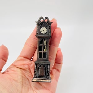 May include: Miniature brown grandfather clock with a white clock face and black hands. The clock has a detailed design with a decorative top and a window showing the pendulum. The clock is held in a person's hand.