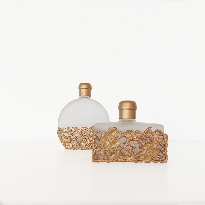 Puede incluir: Dos botellas de perfume de vidrio transparente con detalles decorativos florales dorados. Las botellas est&aacute;n sobre una superficie blanca.