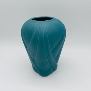 May include: A teal vase with a matte finish. The vase has a rounded shape with a wide opening and a textured, woven-like pattern on the exterior. The design gives the vase a unique, artistic look.