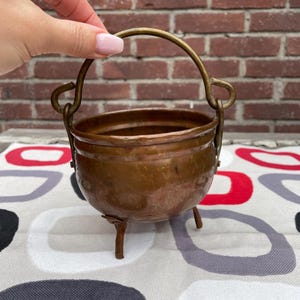 Könnte beinhalten: Ein kleiner, antiker Kupferkessel mit einem Messinggriff und drei kleinen Beinen. Der Kessel hat einen abgerundeten Körper und eine angelaufene Oberfläche, die auf ein hohes Alter hindeutet. Der Griff ist gewölbt und die Beine sind einfach und funktional. Der Kessel wird auf einer gemusterten Oberfläche präsentiert.