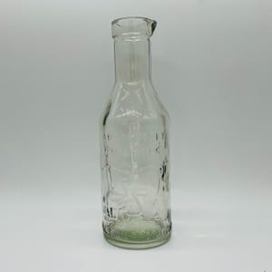Puede incluir: Una botella de vidrio transparente con cuello estrecho y parte superior ligeramente ensanchada. La botella tiene texto en relieve y un diseño de una persona. La botella es probablemente vintage y está vacía.