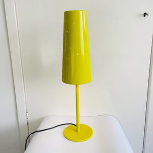 May include: A bright yellow table lamp with a cylindrical shade featuring small, evenly spaced perforations. The lamp has a slender stem and a circular base. The lamp's cord is black. The lamp is on a white surface.