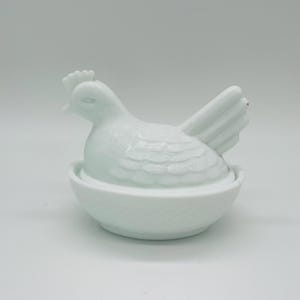 May include: A white ceramic hen figurine sitting in a nest-shaped bowl. The hen has detailed feather and comb features. The bowl has a textured, woven pattern. The figurine is a decorative item.