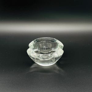 May include: A clear, glass ashtray with a unique, layered design. The ashtray has a circular shape with a textured surface, and is made of transparent glass. The design features two stacked, bowl-shaped sections.
