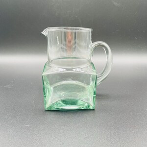 Op de afbeelding: Een heldere glazen kan met een handvat en tuit staat op een vierkante, groen getinte glazen voet. De kan en de voet hebben een geometrisch ontwerp, wat een moderne esthetiek creëert. De kan is leeg en klaar voor gebruik.