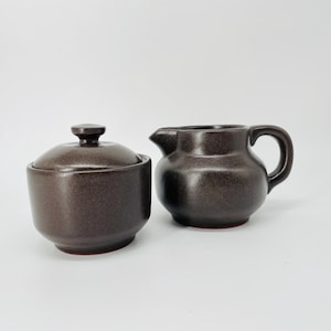 May include: A brown ceramic sugar bowl with a lid and a matching creamer. The sugar bowl is round with a small knob on the lid. The creamer has a handle and a spout. Both pieces have a speckled texture and a reddish-brown base.