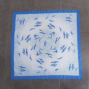 Op de afbeelding: Een blauwe zijden sjaal met een witte achtergrond en een patroon van blauwe vogels. De vogels zijn in een cirkelvormig patroon gerangschikt en kijken allemaal in dezelfde richting.