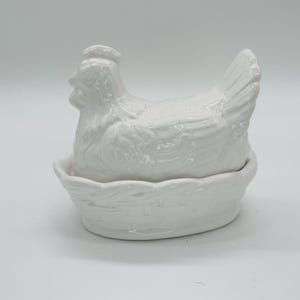 Peut inclure: Un plat en céramique blanche en forme de poule avec un couvercle, conçu pour ressembler à une poule assise dans un nid. Le plat est en céramique brillante et convient à la cuisine ou à la décoration intérieure.