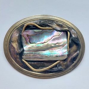 May include: A silver oval-shaped frame with a gold rope design around the edge. Inside the frame is a piece of iridescent abalone shell with a black leather background.