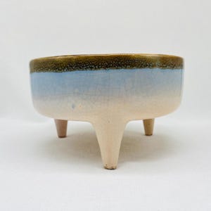 May include: A ceramic planter with a three-legged base. The planter features a light blue and cream color scheme with a brown rim. The planter is designed with a crackle glaze finish.