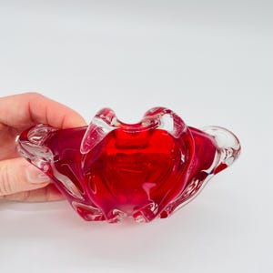 May include: A vibrant red glass bowl with a unique, organic shape. The bowl has a glossy finish and a clear rim, creating a striking contrast. The design features flowing curves and a central indentation, perfect for decorative use.