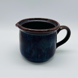 Può includere: Brocca in ceramica blu scuro con bordo e manico marroni. La brocca ha un beccuccio per versare. Questo pezzo artigianale è ideale per servire bevande. Dimensioni: circa 10 cm di altezza.