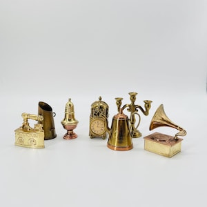 May include: A collection of miniature brass and copper decorative items, including an iron, a coal scuttle, a bell, a clock, a teapot, a candelabra, and a gramophone. The items are arranged on a white surface.