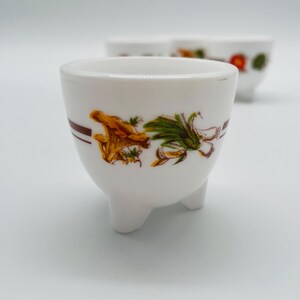 May include: White ceramic egg cups with three small feet. Each cup has a brown stripe and a colourful floral design with orange, green, and yellow accents. Several cups are visible, with one in sharp focus.