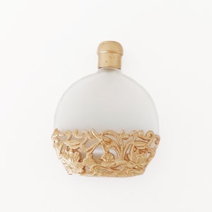 Puede incluir: Un frasco de perfume de vidrio esmerilado con un dise&ntilde;o de filigrana dorada alrededor de la base. El frasco tiene una tapa dorada.