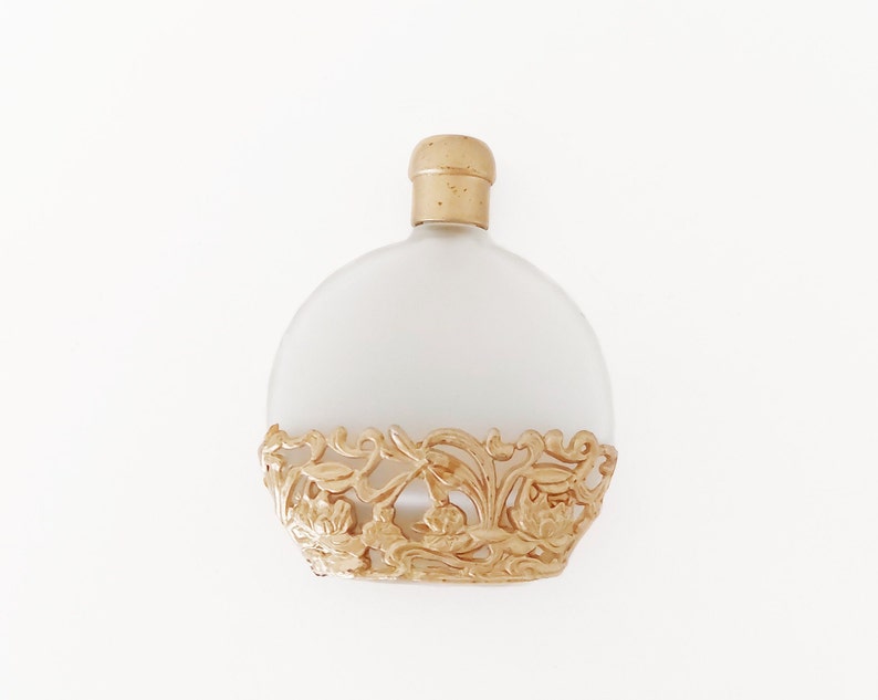 Puede incluir: Un frasco de perfume de vidrio esmerilado con un dise&ntilde;o de filigrana dorada alrededor de la base. El frasco tiene una tapa dorada.