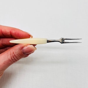 Peut inclure: Petite fourchette de service ancienne avec un manche crème et une fourchette argentée à deux dents. Le manche est lisse et effilé, et les dents de la fourchette sont fines. La fourchette est tenue dans une main sur fond blanc.