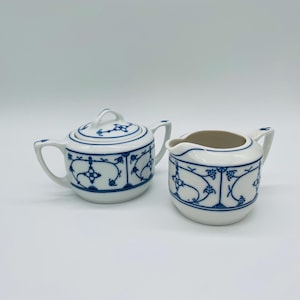 May include: A white ceramic sugar bowl with a lid and a creamer. Both pieces feature a blue floral and vine pattern. The sugar bowl has two handles and a small handle on the lid. The creamer has a handle and a spout.