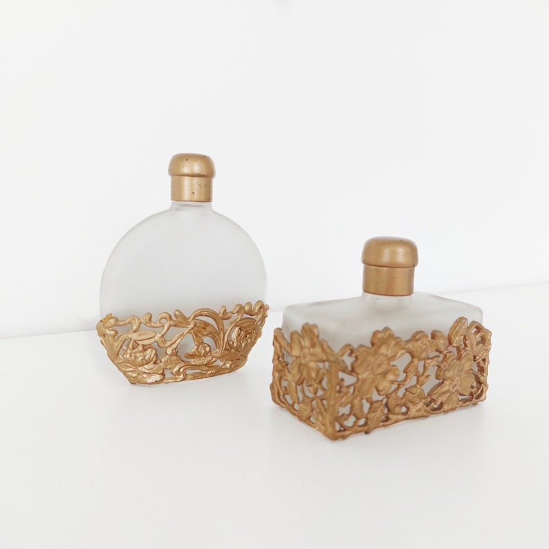 Puede incluir: Dos botellas de perfume de vidrio esmerilado con detalles de filigrana dorada. Las botellas est&aacute;n vac&iacute;as y tienen una est&eacute;tica vintage.