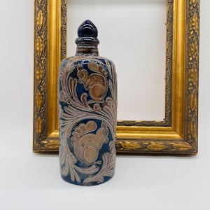 Può includere: Bottiglia decorativa in ceramica con smalto blu scuro e un design dettagliato con scoiattoli e motivi a vortice in argento e marrone. La bottiglia ha un tappo blu scuro.
