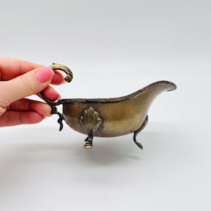 Könnte beinhalten: Eine Vintage-Sauciere in Bronzetönen mit aufwendigen Details. Das Boot hat einen geschwungenen Griff, ein muschelartiges Design an der Seite und drei zarte Füße. Das Design ist elegant und von antiken Vorbildern inspiriert.