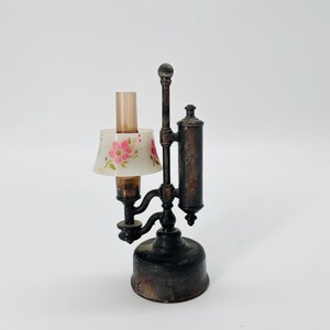 Pode incluir: Lâmpada antiga em miniatura com abajur floral. A lâmpada tem uma base de metal escuro e um reservatório cilíndrico. O abajur é branco com flores cor-de-rosa e folhas verdes. É um objeto decorativo.