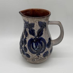 Könnte beinhalten: Ein Keramikkrug mit braunem Innenraum und grauem Äußeren. Der Krug ist mit blauen Obst- und Blattmotiven verziert. Er hat einen Henkel und einen Ausguss zum Ausgießen.